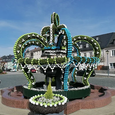Brunnen mit grünen, weißen und blauen Ostereiern und Pflanzen in einem Dorfplatz bei Sonnenschein.