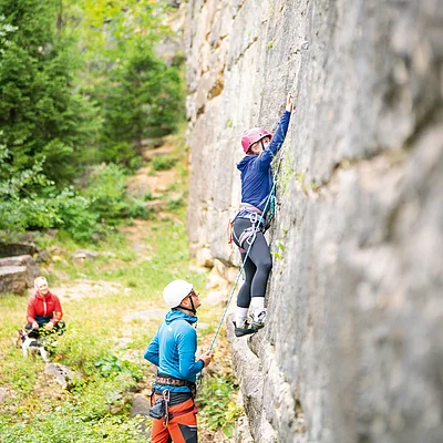 Frau klettert an Felswand, Mann sichert sie mit Seil, weitere Person sitzt im Hintergrund auf Wiese.