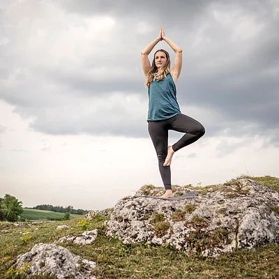 Frau macht Yoga im Baum-Pose auf Felsen mit bewölktem Himmel und grüner Landschaft im Hintergrund.