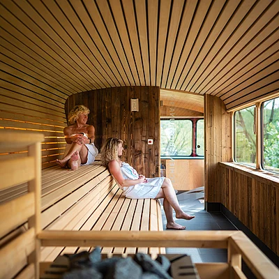 Zwei Frauen in weißen Handtüchern sitzen in einer Holzsauna mit großen Fenstern und Saunaofen im Vordergrund.