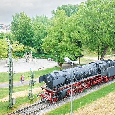 Dampflokomotive auf Gleis neben Park mit Spielplatz und Spaziergängern bei bewölktem Himmel