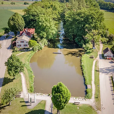 Luftaufnahme eines rechteckigen Teichs mit Bäumen, Wegen und Häusern an einem sonnigen Tag