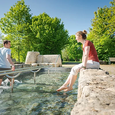 Mann watet im Wasserbecken, Frau sitzt am Beckenrand und taucht Füße ins Wasser, Park mit Bäumen im Hintergrund.