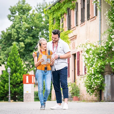 Paar mit Audioguides liest Stadtplan vor Gebäude mit Efeu und Informationsschild im Hintergrund.