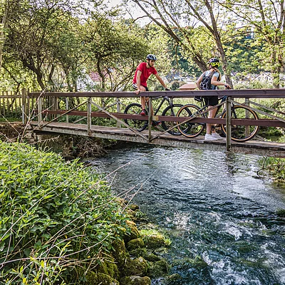 Zwei Radfahrer mit Helm schieben ihre Fahrräder über eine schmale Brücke über einen Bach in bewaldeter Landschaft.