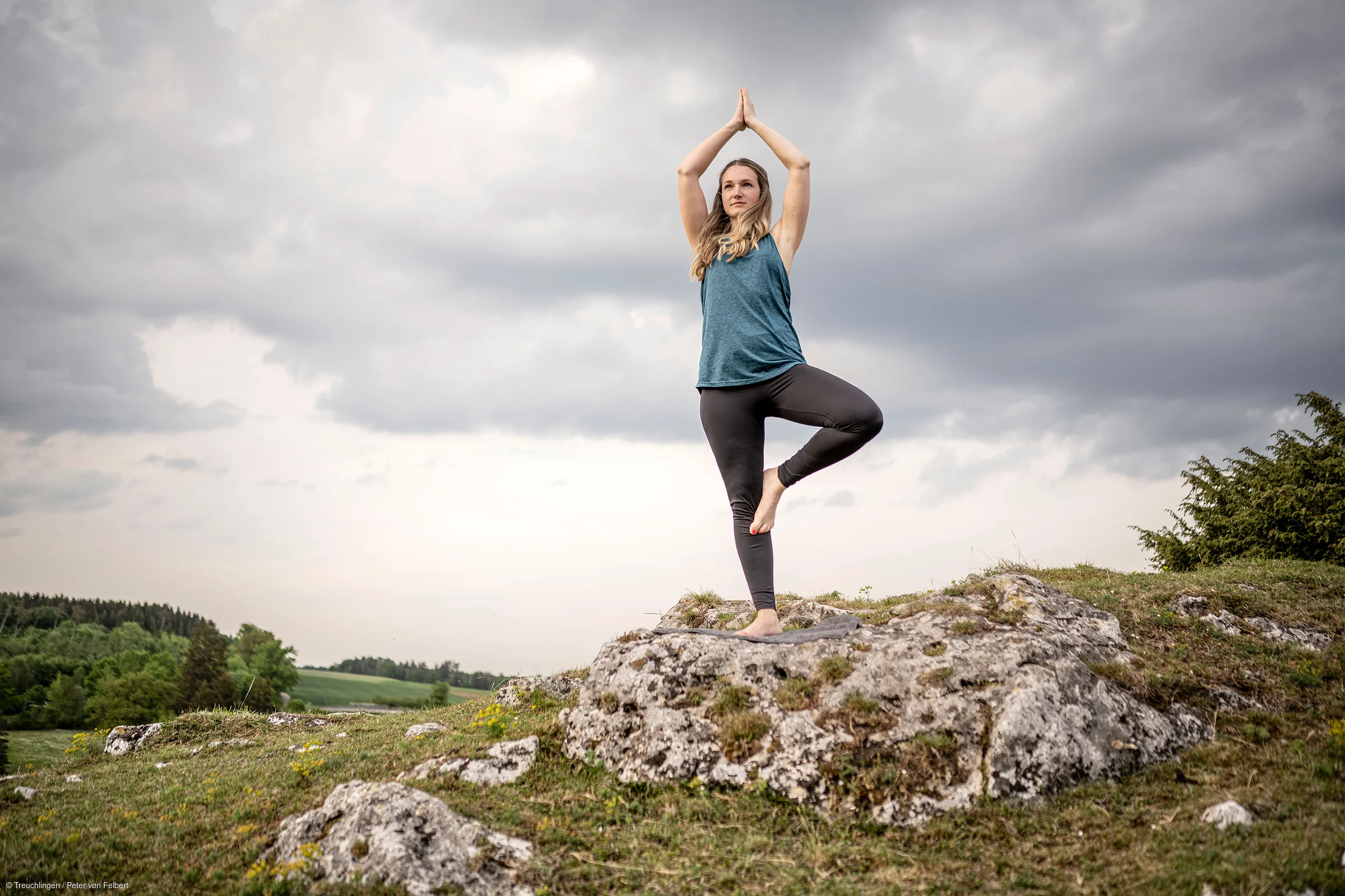 Frau macht Yoga im Baum-Pose auf Felsen mit bewölktem Himmel und grüner Landschaft im Hintergrund.