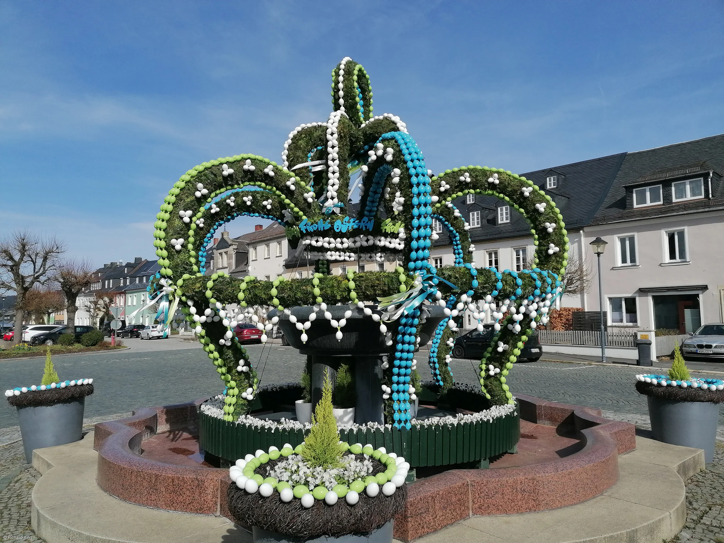 Brunnen mit grünen, weißen und blauen Ostereiern und Pflanzen in einem Dorfplatz bei Sonnenschein.