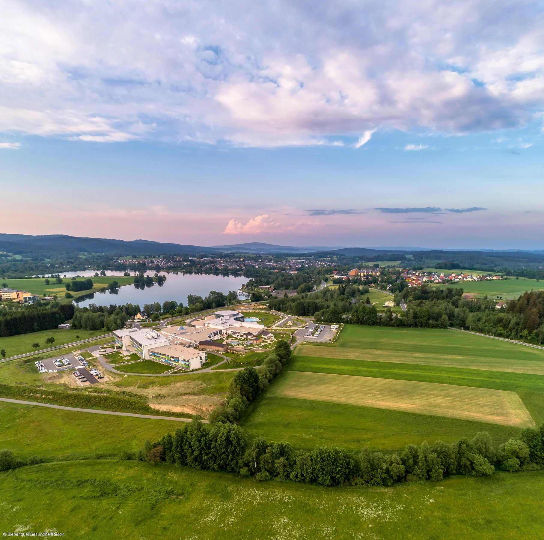 Luftaufnahme von Feldern, einem See und einem Gebäudekomplex unter bewölktem Himmel bei Abendlicht