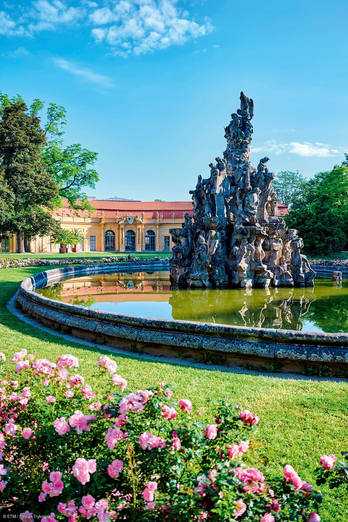 Brunnen mit Figuren in einem runden Wasserbecken vor einem gelben Schloss und blühenden Rosen im Park bei blauem Himmel