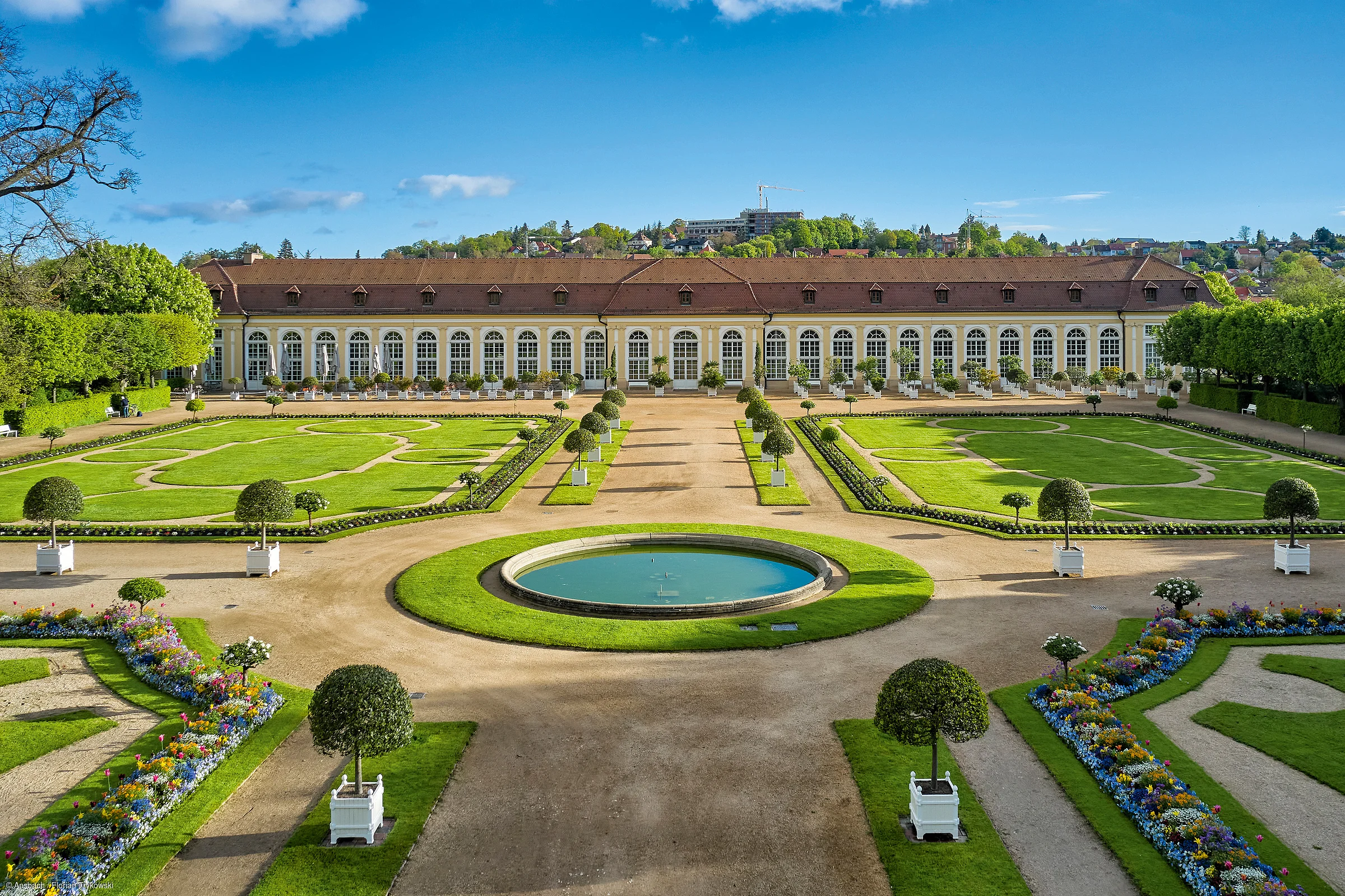 Barocker Garten mit symmetrischen Wegen, Blumenbeeten, Büschen und einem großen Gebäude im Hintergrund bei blauem Himmel.