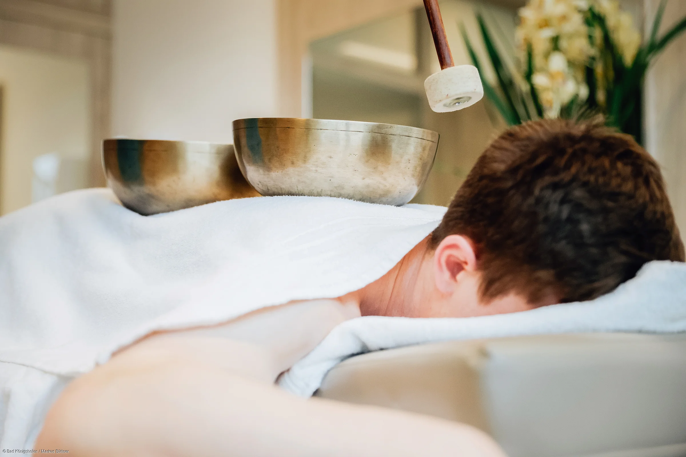 Person liegt mit Kopf nach unten auf Massageliege, Klangschalen und Schlägel auf Rücken, Pflanzen im Hintergrund