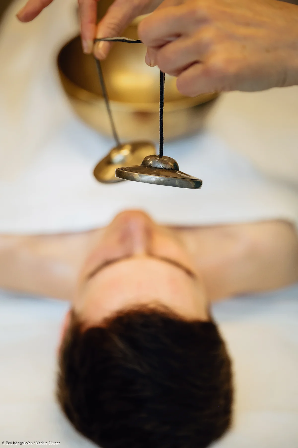 Person liegt mit geschlossenen Augen, darüber werden kleine Klangschalen an Schnüren gehalten.