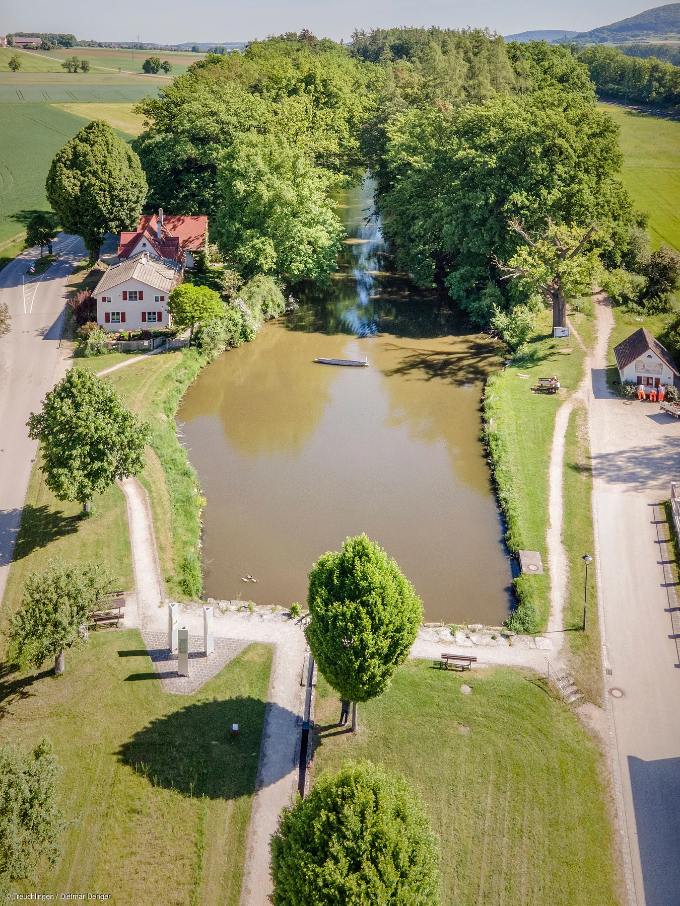 Luftaufnahme eines rechteckigen Teichs mit Bäumen, Wegen und Häusern an einem sonnigen Tag