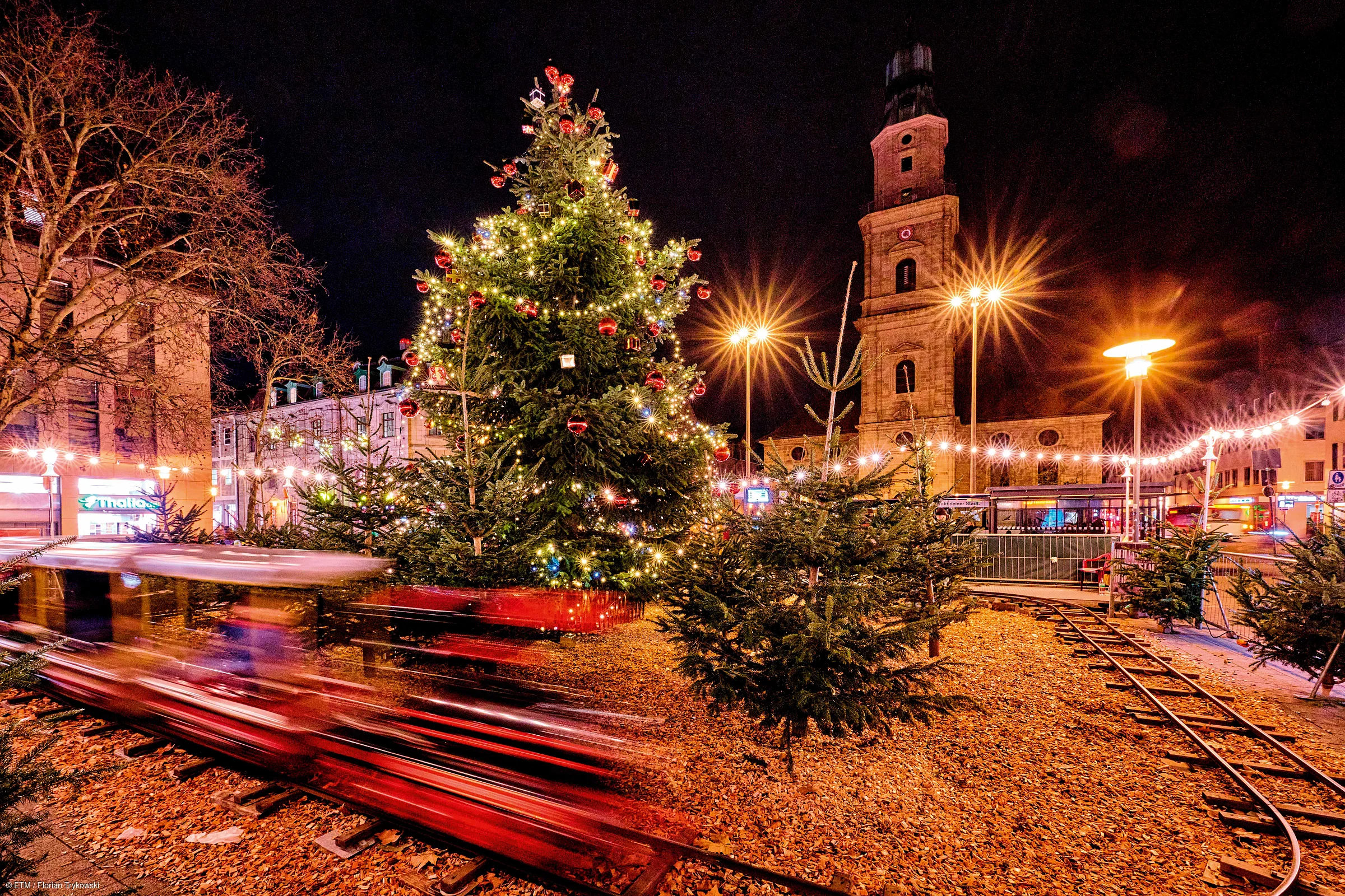 Weihnachtsmarkt bei Nacht mit beleuchtetem Weihnachtsbaum, Mini-Zug und historischem Turm im Hintergrund.