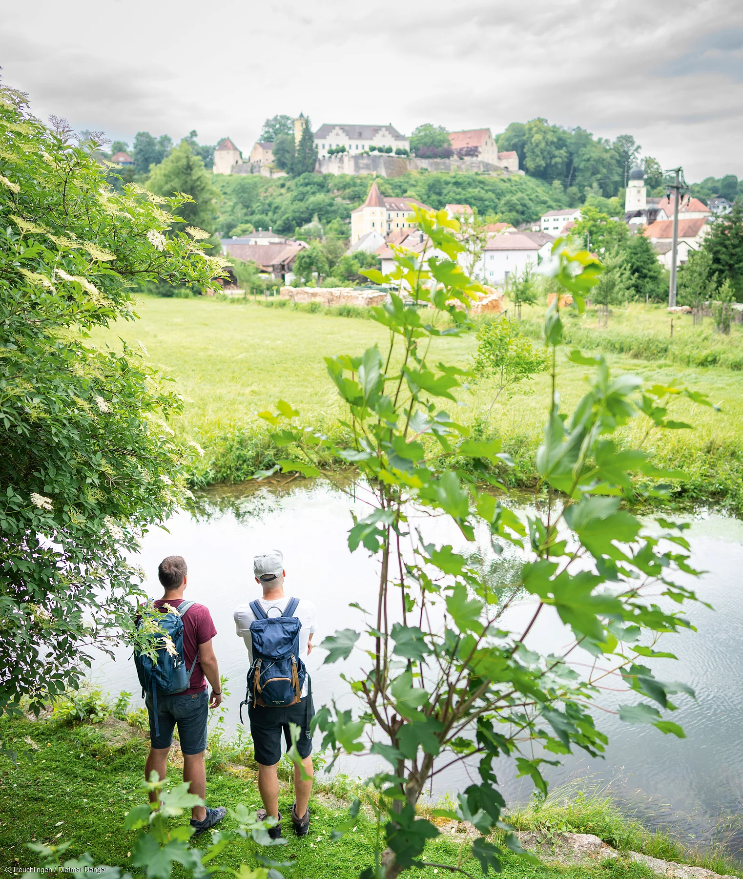 Zwei Männer mit Rucksäcken stehen an einem Teich, im Hintergrund Dorf und Burg auf bewaldetem Hügel.