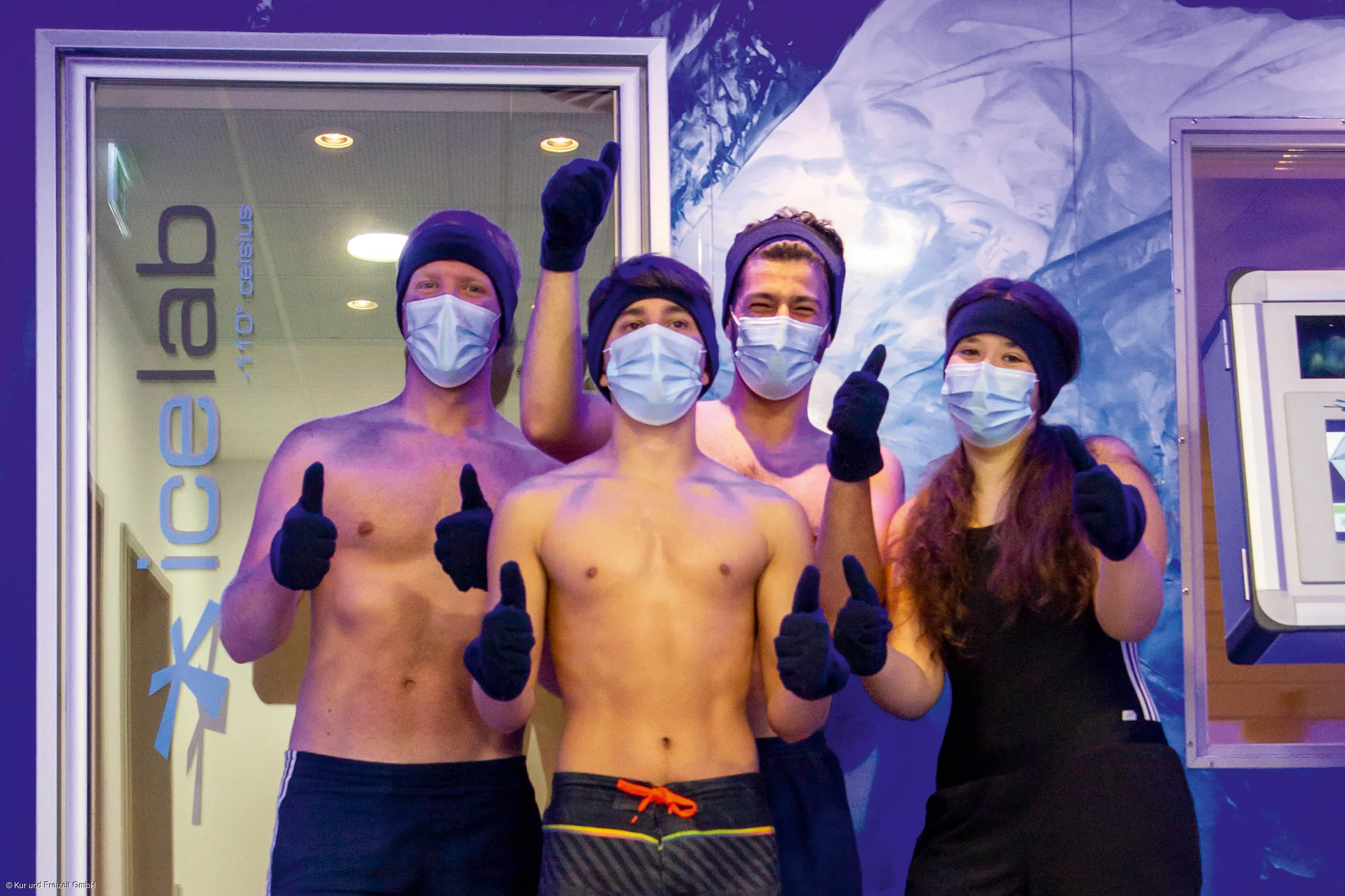 Vier Personen mit Mundschutz, Mütze und Handschuhen zeigen Daumen hoch vor einem Eisraum mit der Aufschrift „icelab -110° Celsius“.