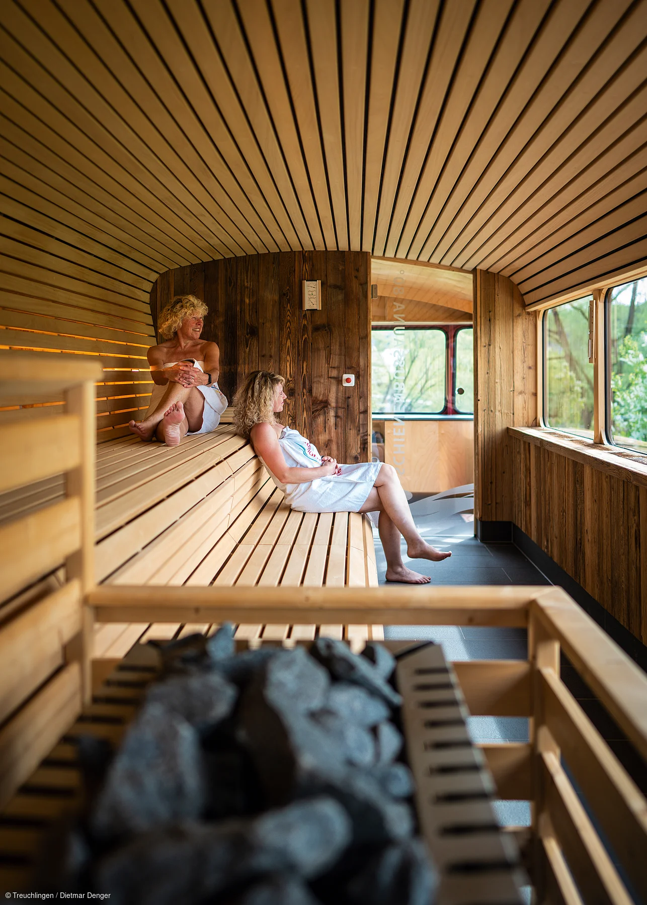Zwei Frauen in weißen Handtüchern sitzen in einer Holzsauna mit großen Fenstern und Saunaofen im Vordergrund.