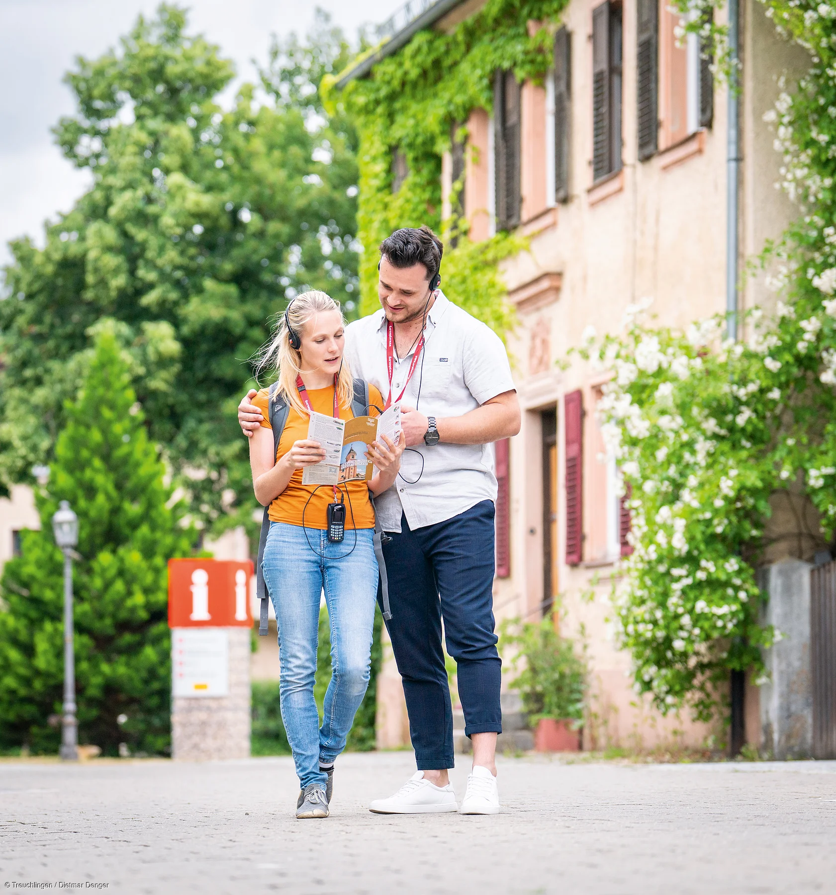 Paar mit Audioguides liest Stadtplan vor Gebäude mit Efeu und Informationsschild im Hintergrund.