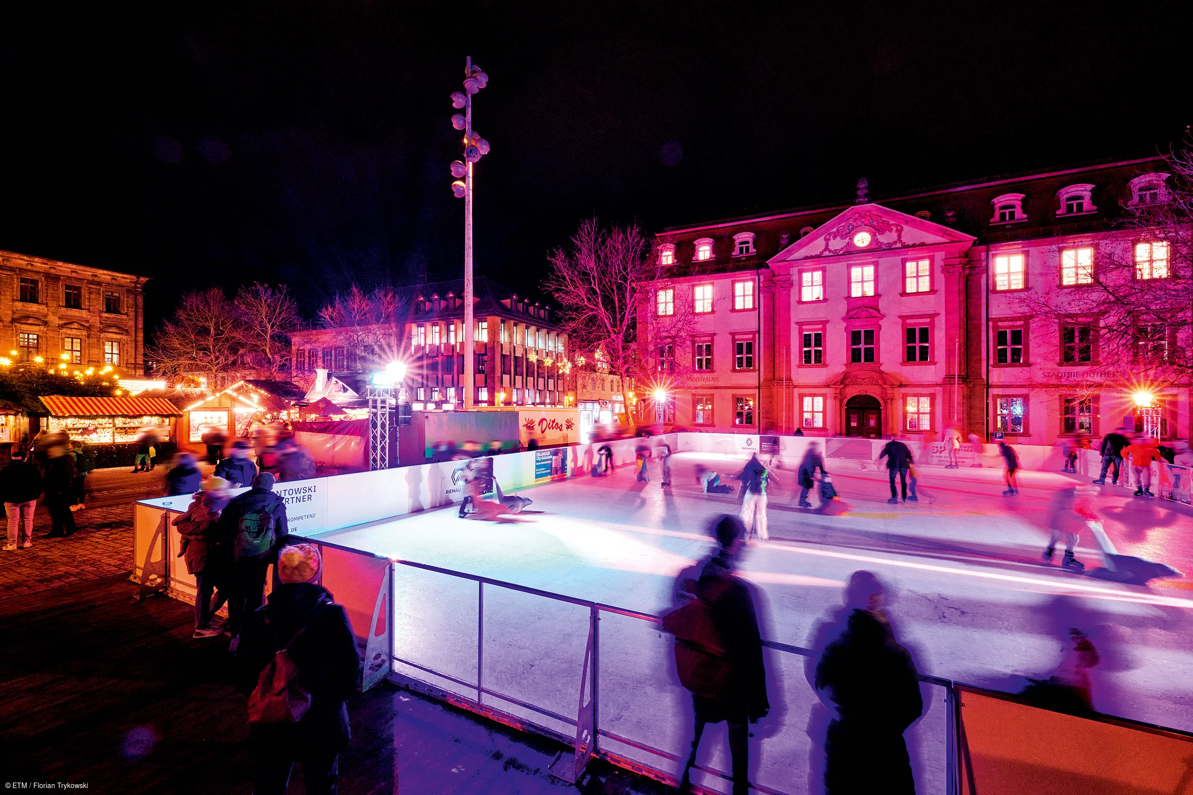 Eisbahn mit mehreren Eisläufern und beleuchteten Gebäuden bei Nacht in einer Stadt.
