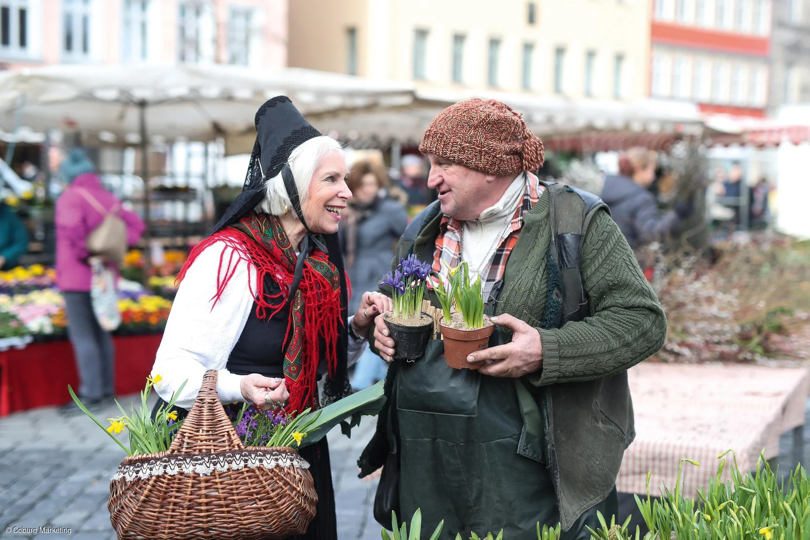 Ältere Frau in Tracht mit Korb und älterer Mann mit Mütze tauschen sich auf einem Blumenmarkt aus.