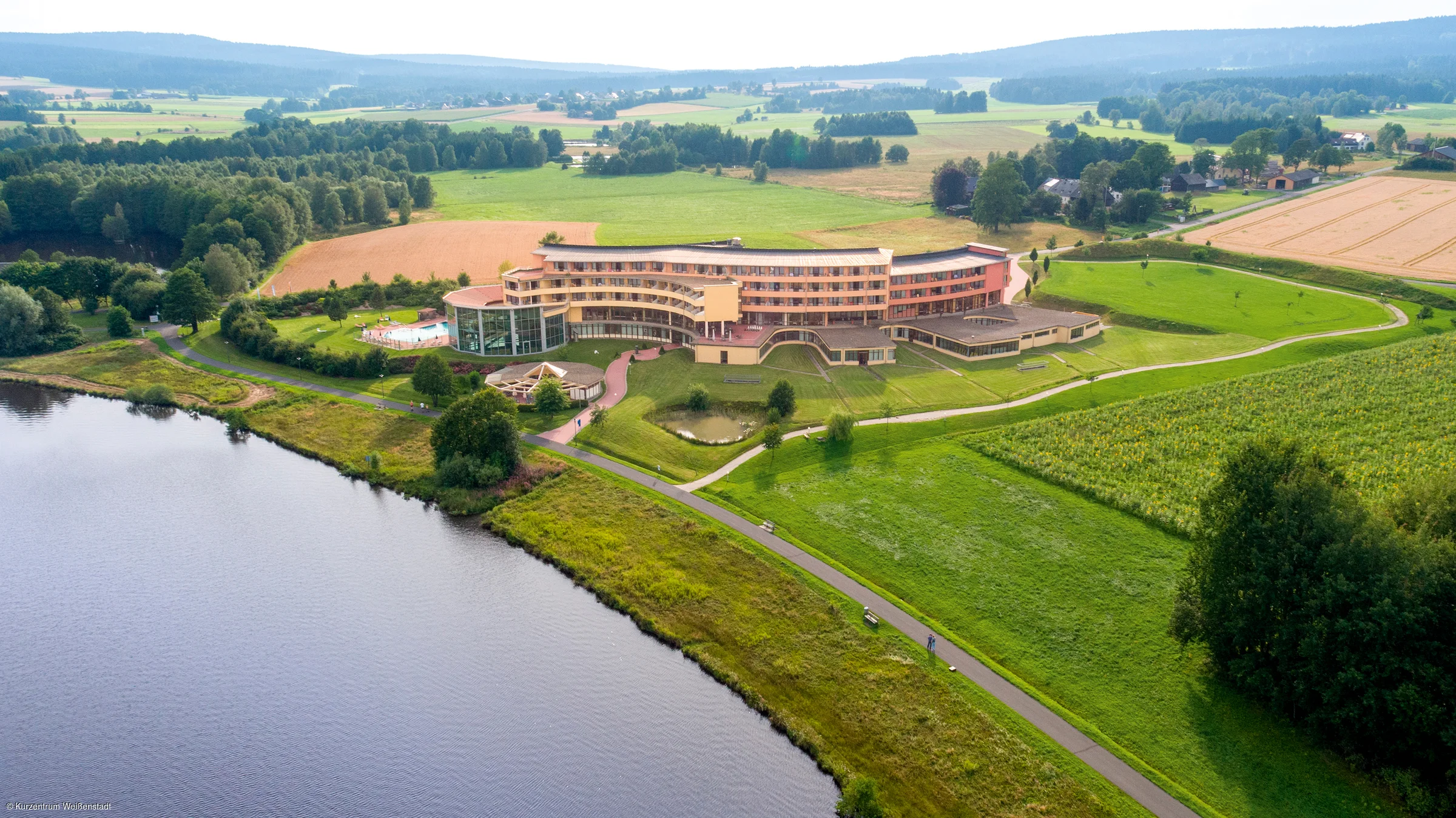 Luftaufnahme eines großen Gebäudes an einem Seeufer, umgeben von Wiesen, Feldern und Bäumen.