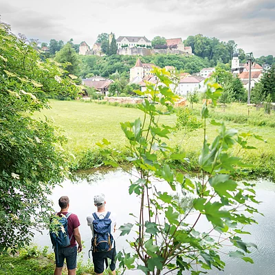 Zwei Männer mit Rucksäcken stehen an einem Teich, im Hintergrund Dorf und Burg auf bewaldetem Hügel.