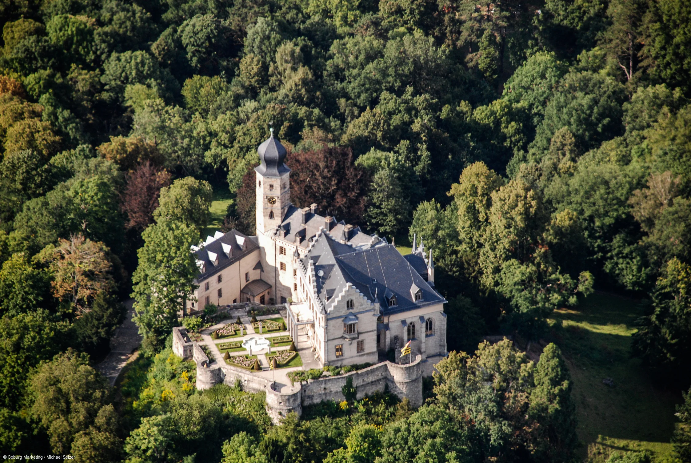 Luftaufnahme eines Schlosses mit Turm und Garten, umgeben von dichtem Wald.