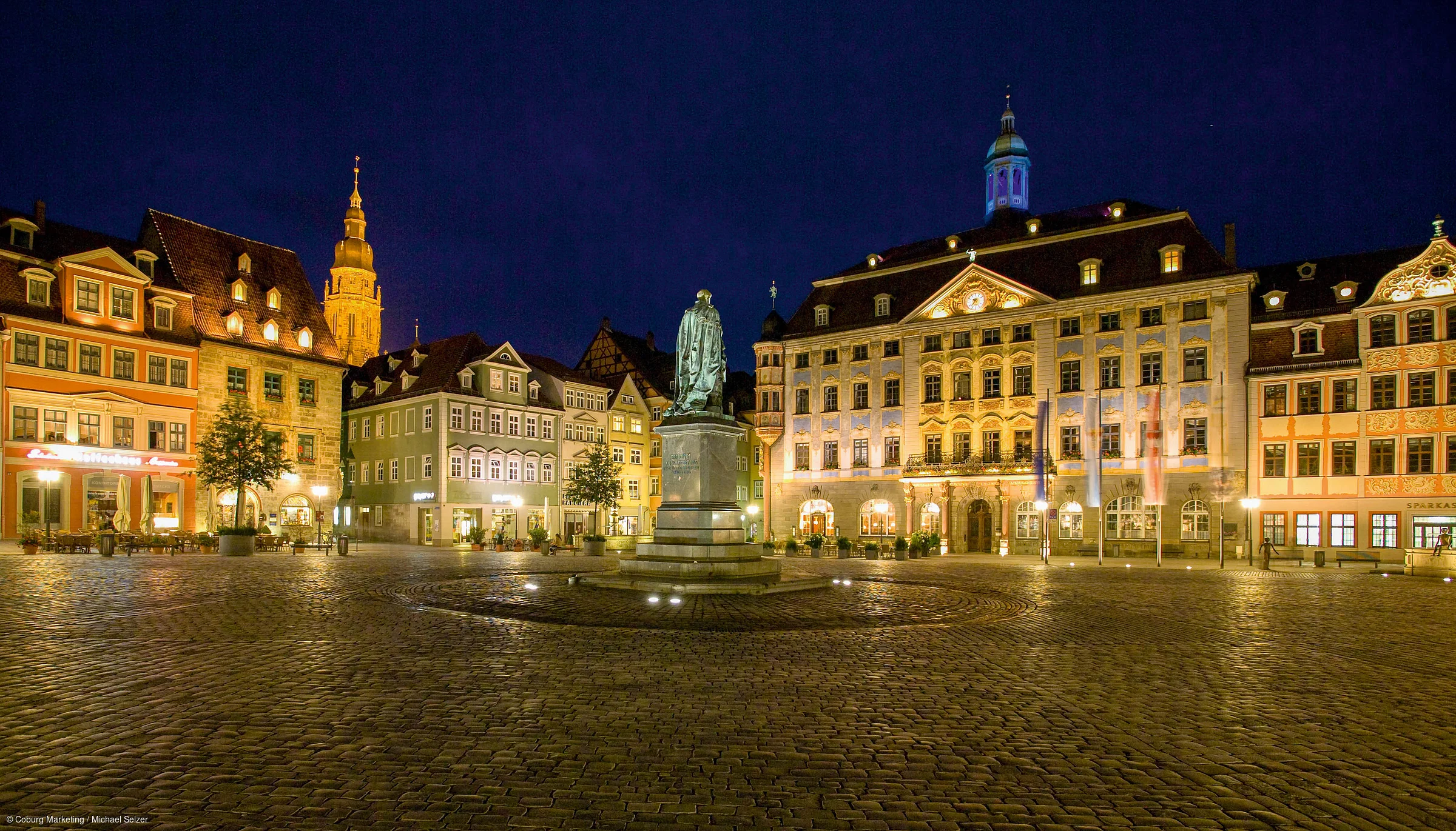 Beleuchteter Marktplatz mit Statue und historischen Gebäuden bei Nacht, Kopfsteinpflaster sichtbar.