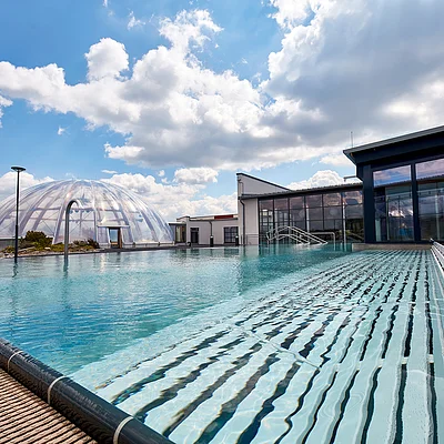 Außenpool mit klar blauem Wasser, transparentem Kuppelgebäude und moderner Architektur bei bewölktem Himmel.