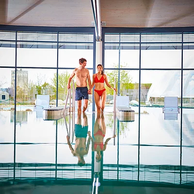 Paar in Badebekleidung steigt an Geländern aus einem Hallenbad mit großen Fenstern und Liegestühlen.