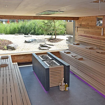 Innenraum einer Sauna mit Holzbänken, zentralem Ofen mit Steinen und großem Fenster zum Gartenbereich