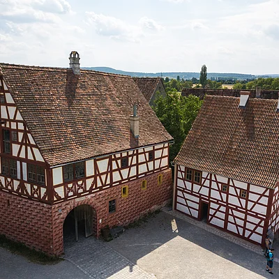 Zwei Fachwerkhäuser mit roten Ziegeldächern und Menschen auf gepflastertem Hof bei bewölktem Himmel.