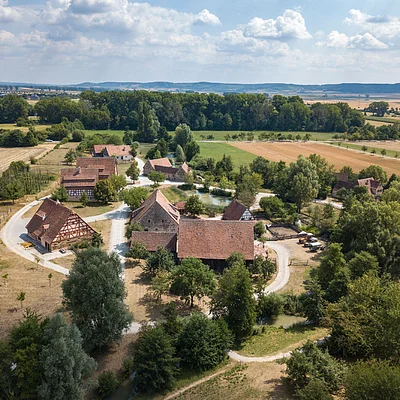 Luftaufnahme eines ländlichen Dorfes mit Fachwerkhäusern, Feldern und Bäumen unter bewölktem Himmel.