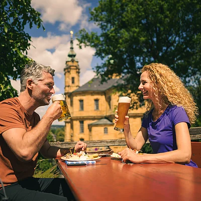 Mann und Frau sitzen draußen an Holztisch und trinken Bier vor historischer Kirche bei Sonnenschein.