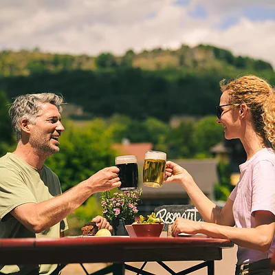 Mann und Frau stoßen mit Biergläsern an, sitzen draußen an einem Tisch mit Pflanzen und Berg im Hintergrund.