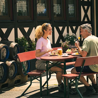Zwei Personen sitzen an einem roten Tisch mit Bänken vor einem Fachwerkhaus und essen im Freien.