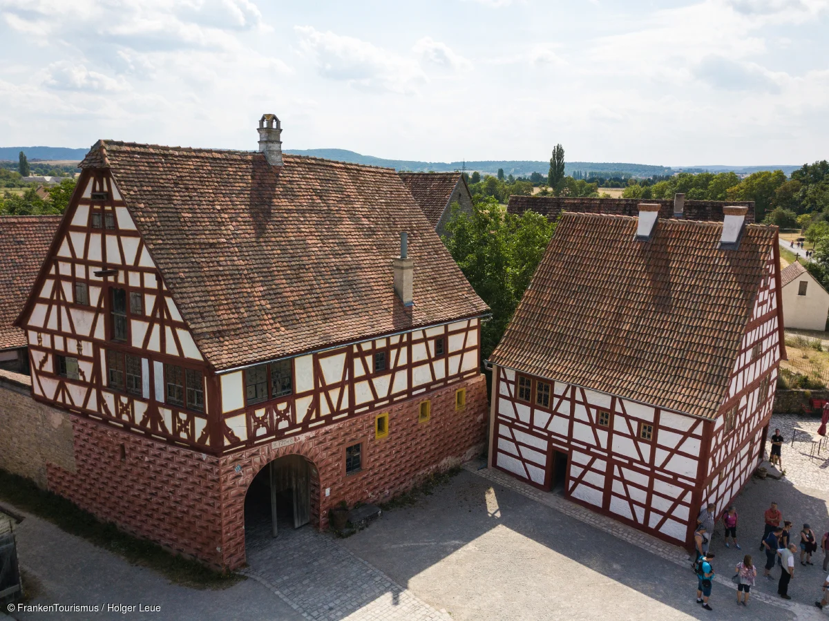Zwei Fachwerkhäuser mit roten Ziegeldächern und Menschen auf gepflastertem Hof bei bewölktem Himmel.