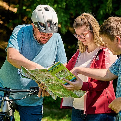 Drei Personen mit Fahrradhelm und Rucksack schauen gemeinsam auf eine Wanderkarte im Wald.
