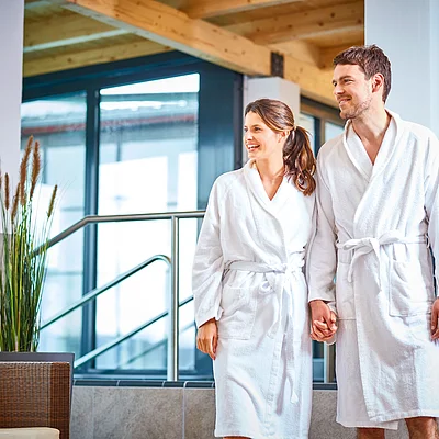 Paar in weißen Bademänteln hält Händchen in einem modernen Wellnessbereich mit großen Fenstern.