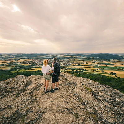 Zwei Wanderer stehen auf einem Felsen und blicken auf eine weite Landschaft mit Feldern und Wäldern.