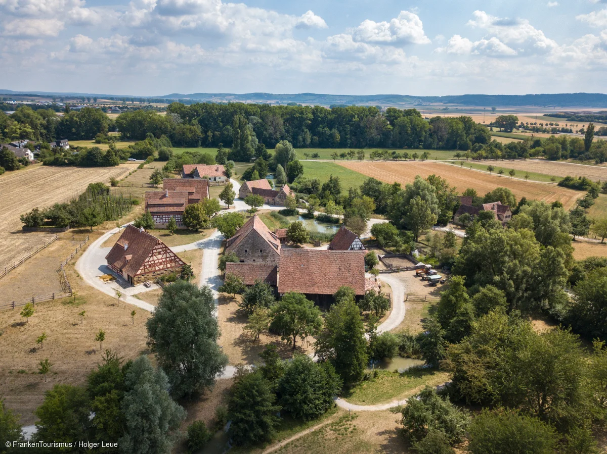 Luftaufnahme eines ländlichen Dorfes mit Fachwerkhäusern, Feldern und Bäumen unter bewölktem Himmel.