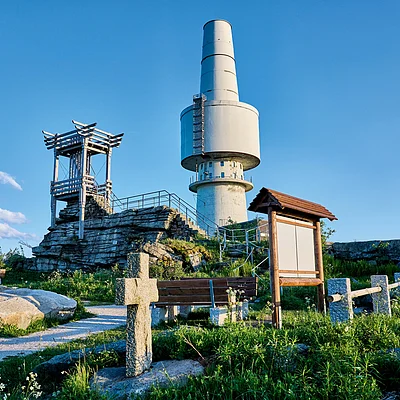 Felsiger Hügel mit Aussichtsturm, Informationsschild, Bank und großem Sendeturm bei klarem Himmel.