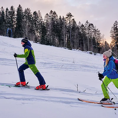 Zwei Personen auf schneebedecktem Hang vor Wald mit Abendhimmel.