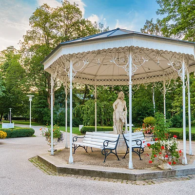 Weißes Pavillon mit Statue und Bänken in einem Park mit Bäumen und Blumenarrangements