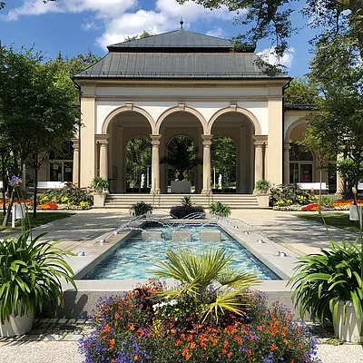 Gebäude mit Arkaden, davor ein Springbrunnen und Blumenbeete in einem Park bei sonnigem Wetter.