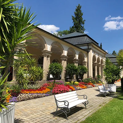 Außenbereich mit weißer Bank, bunten Blumen und Arkaden eines hellen Gebäudes bei Sonnenschein.