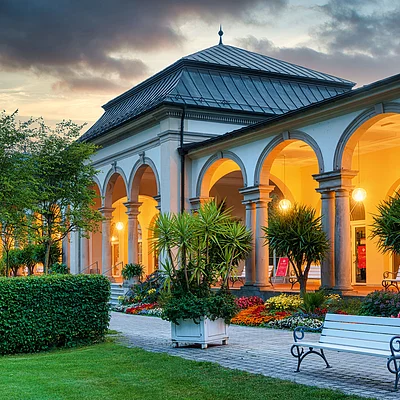 Gebäude mit Arkaden, beleuchteten Bögen, Blumenbeeten, Bänken und Bäumen bei Abenddämmerung.