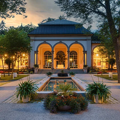 Gebäude mit drei großen Bögen, beleuchtet, davor ein rechteckiger Brunnen und Pflanzen, Abendstimmung im Park.