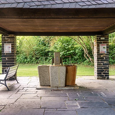 Überdachter Bereich mit drei Wasserbrunnen aus Stein und einer weißen Parkbank, umgeben von Grünflächen.