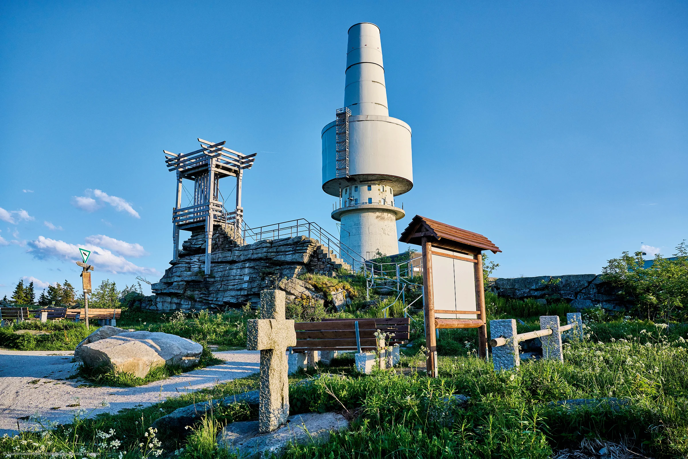 Felsiger Hügel mit Aussichtsturm, Informationsschild, Bank und großem Sendeturm bei klarem Himmel.